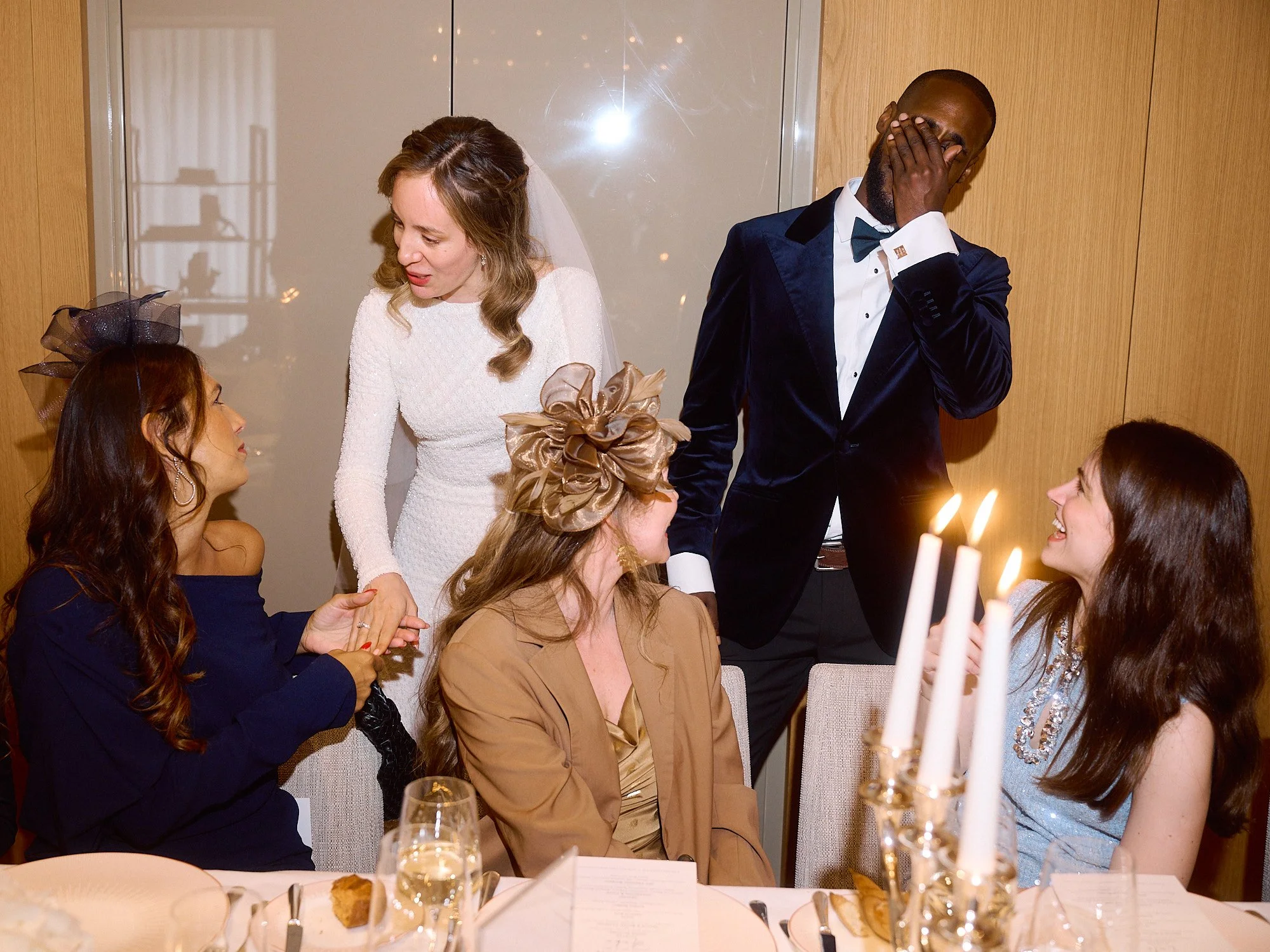 A lively and candid wedding moment captured by Wayne La Photography, showing the bride leaning in to greet her guests at an elegant dinner table while the groom stands beside her laughing with his hand over his face. Warm candlelight and soft reflect