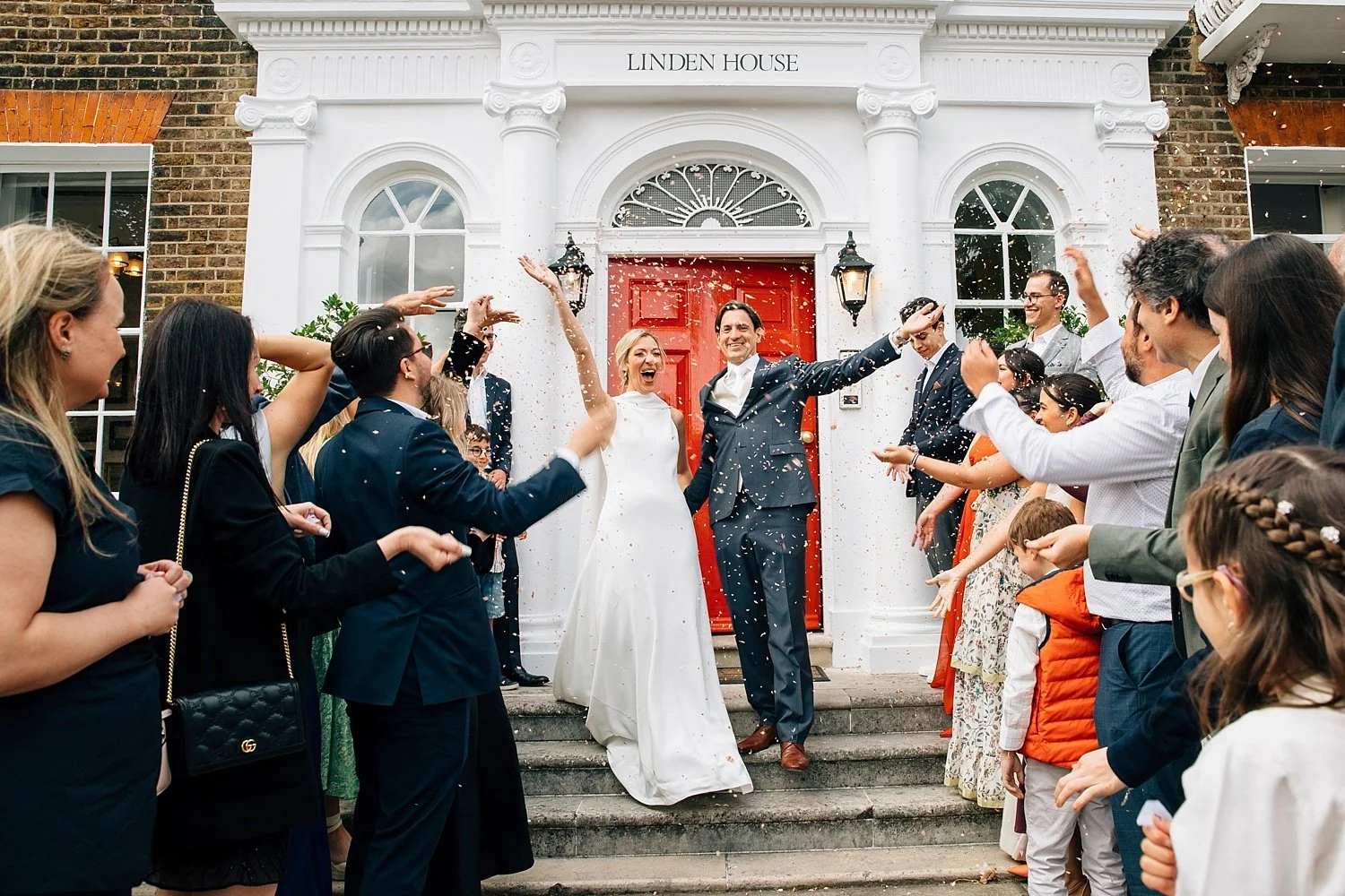 A joyful confetti exit outside Linden House captures a newly-married couple laughing and celebrating beneath the venue’s iconic red door. Surrounded by cheering guests, the pair hold hands and throw their arms up as petals fill the air. This vibrant 