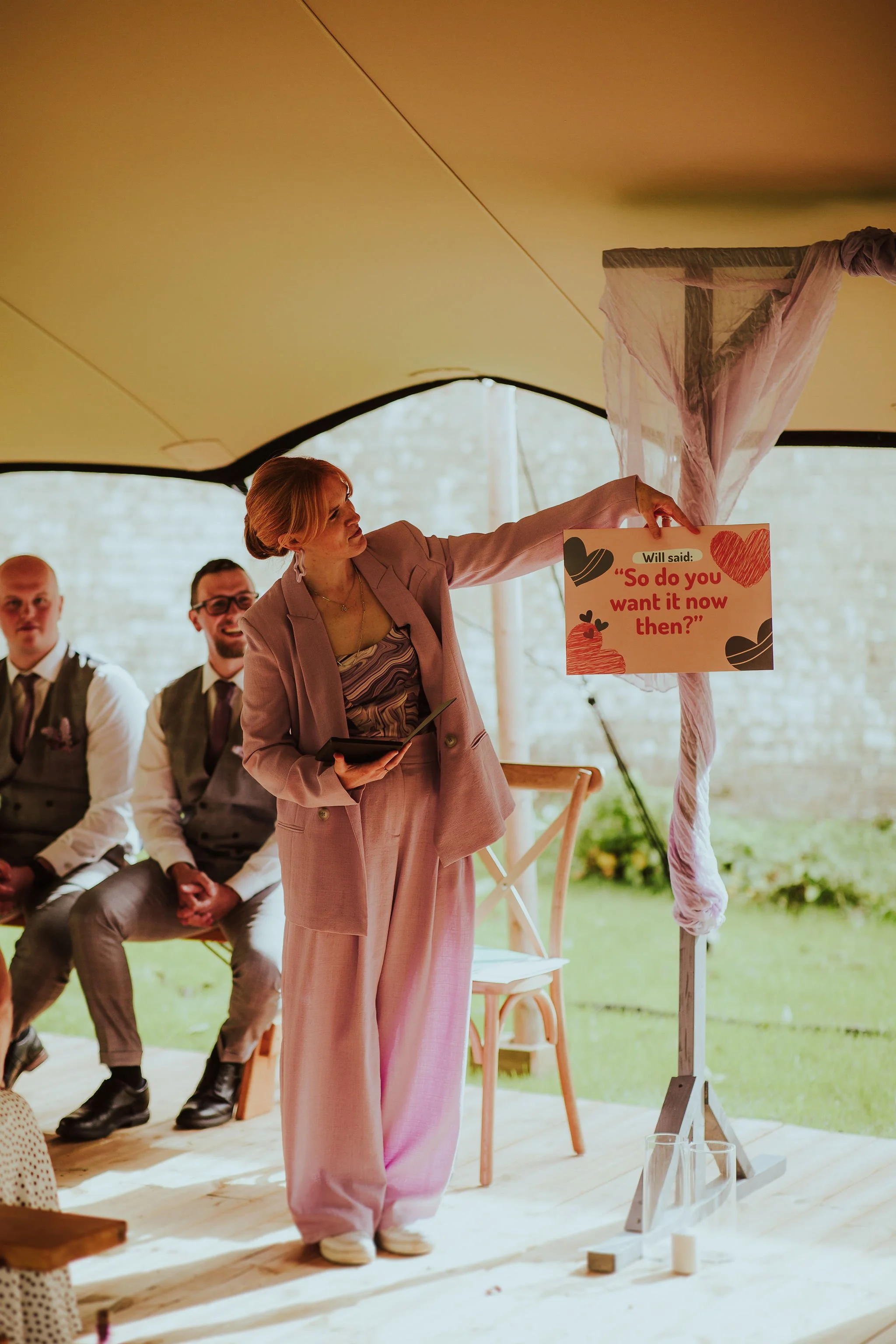 Wedding celebrant Meg Senior leading a joyful outdoor ceremony under a tipi-style canopy. The couple and guests share laughter surrounded by colourful paper fans and summer flowers, creating a relaxed and vibrant wedding atmosphere. Photrto by Salsab