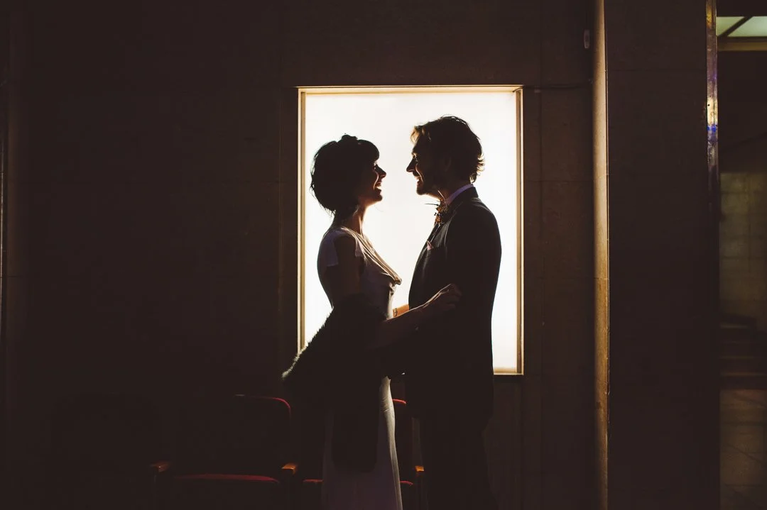 A dramatic and intimate wedding portrait captured by Barbara K Photography, showing a couple standing in silhouette against a softly glowing backlit panel. Their faces are close as they share a quiet moment, creating a romantic contrast between the d