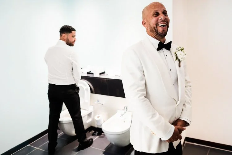 A brilliantly candid and humorous moment captured by Matt Parry Photography, showing two men getting ready before a wedding. One man stands in the foreground wearing a sharp white tuxedo jacket with a black bow tie and boutonniere, laughing openly wi