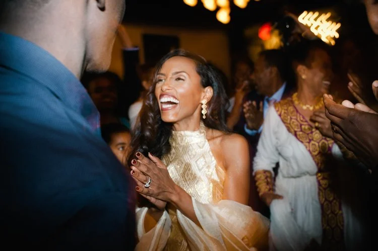 A joyful, high-energy moment on the dance floor as the bride laughs and celebrates with guests around her. Dressed in a shimmering gold gown and glowing with happiness.