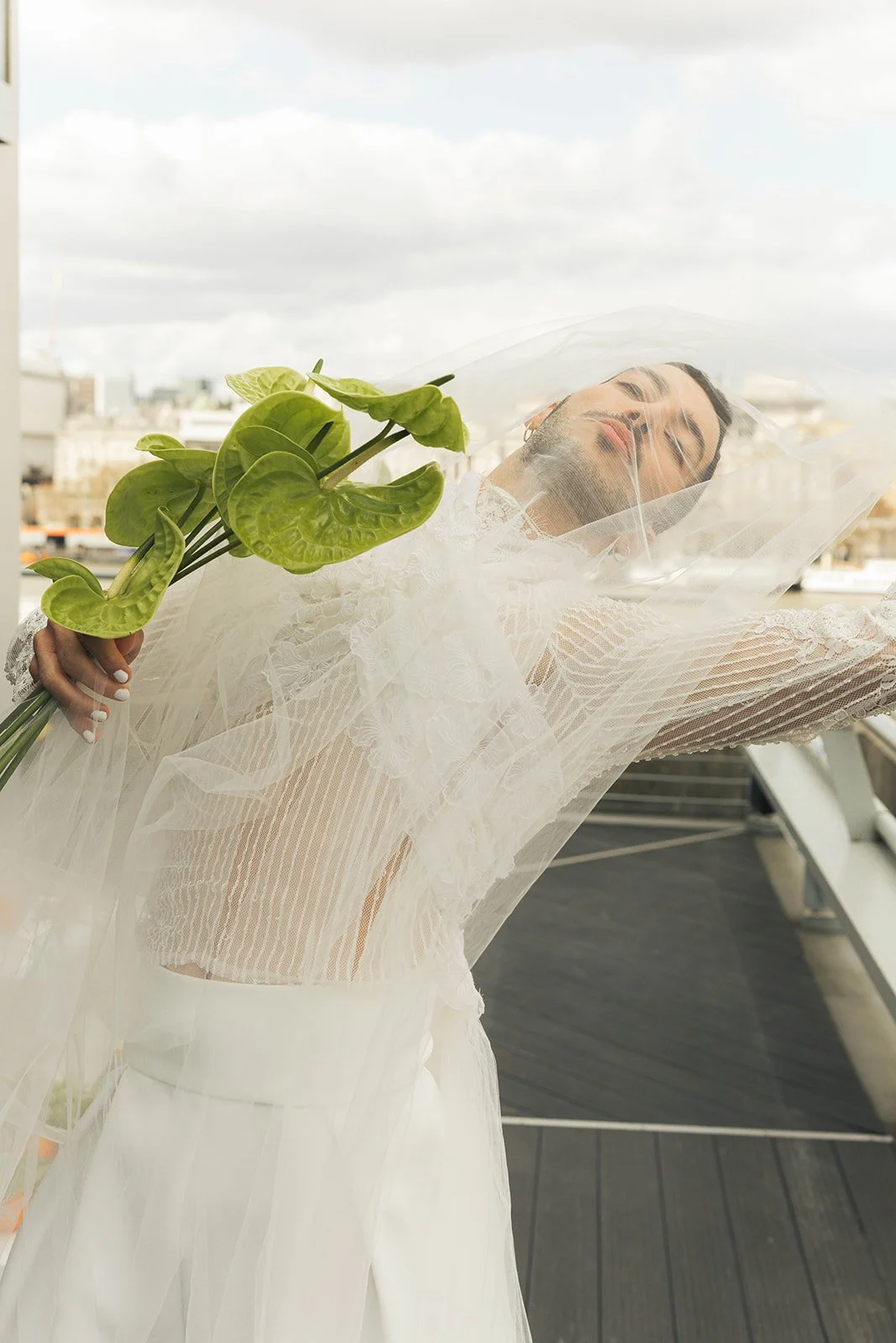 A person stands outdoors, leaning back with their head tilted and eyes closed, wrapped in layers of sheer white fabric. One arm extends outward while the other holds a small bouquet of green, heart-shaped leaves. The veil and textured fabric catch th