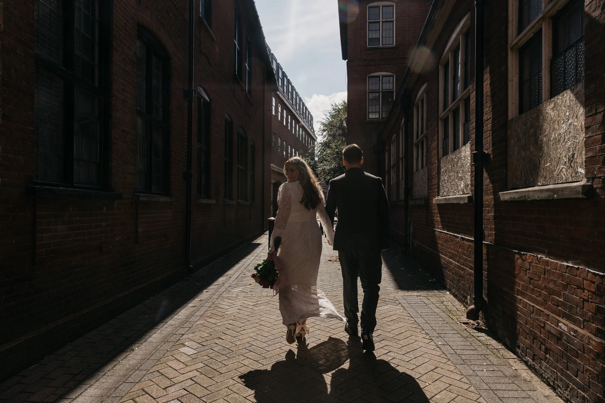 A cinematic and atmospheric wedding moment captured by YU Photography, showing the couple walking hand in hand through a narrow brick alleyway as golden sunlight streams in behind them. The bride glances back over her shoulder, her lace gown catching