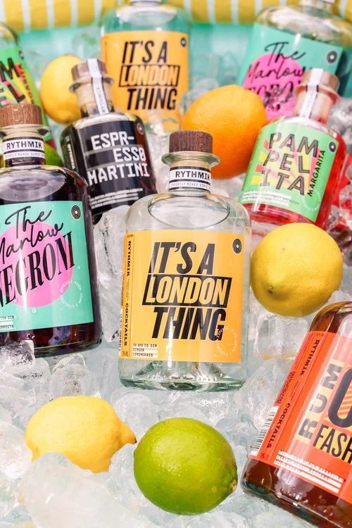 A vibrant display of Rythmik Cocktails bottles chilling on a bed of ice, surrounded by fresh lemons and limes. Featured flavours include It’s A London Thing, The Marlow Negroni, Pam Pella Margarita, Espresso Martini, and Rum Old Fashioned. The colour