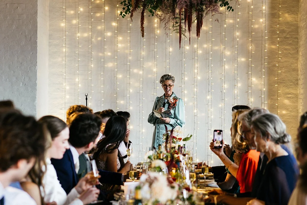 Arabella stands alongside the couple for a posed portrait, all three facing the camera and smiling. The bride holds a large bouquet of mixed flowers, while the celebrant and partner stand close on either side, framed by a brick wall backdrop in natur