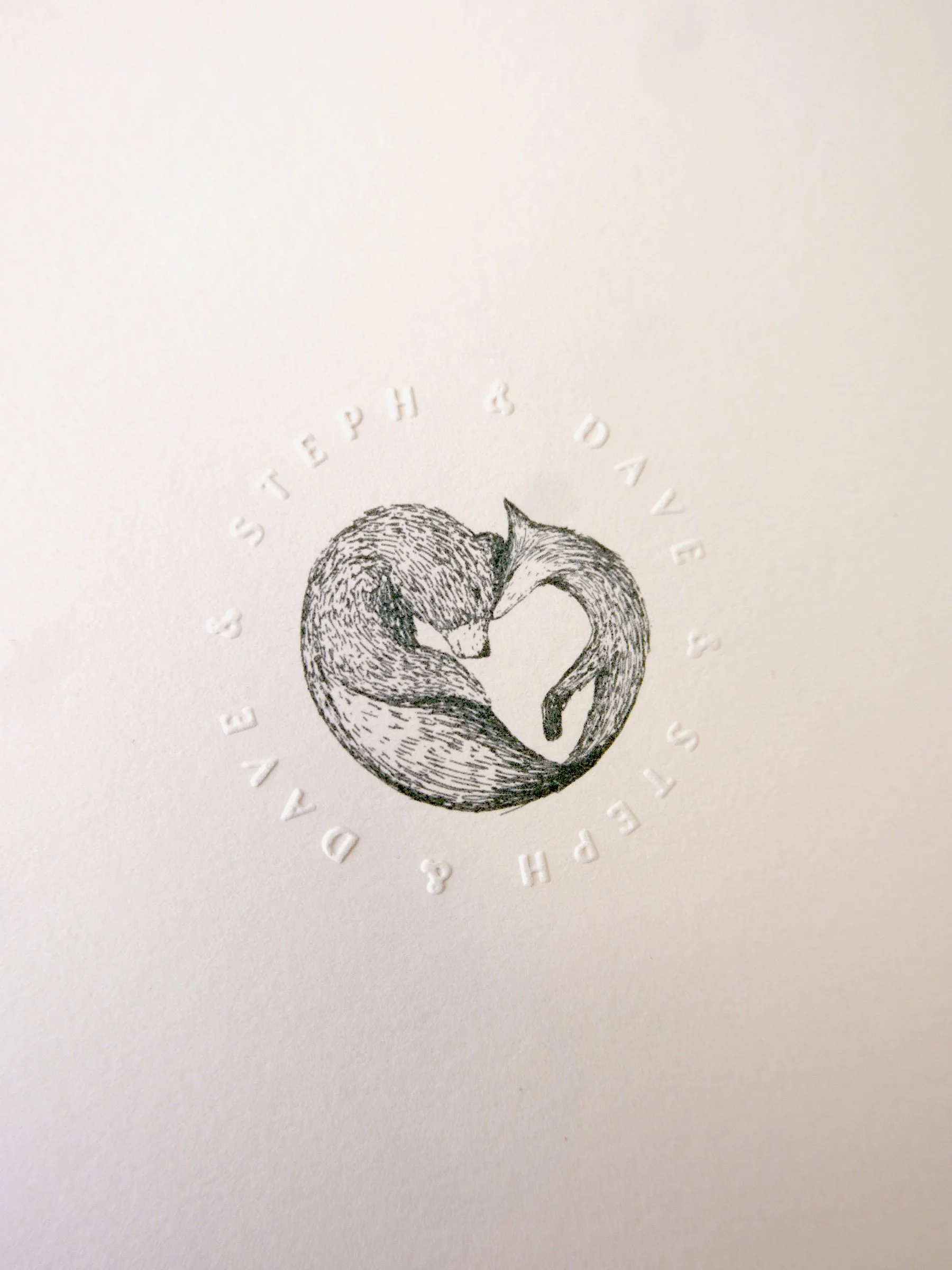 A delicate wedding stationery detail featuring an intricately illustrated pair of foxes curled into a heart shape with the couple’s names embossed in a circular design on soft textured card creating a romantic woodland inspired motif ideal for nature