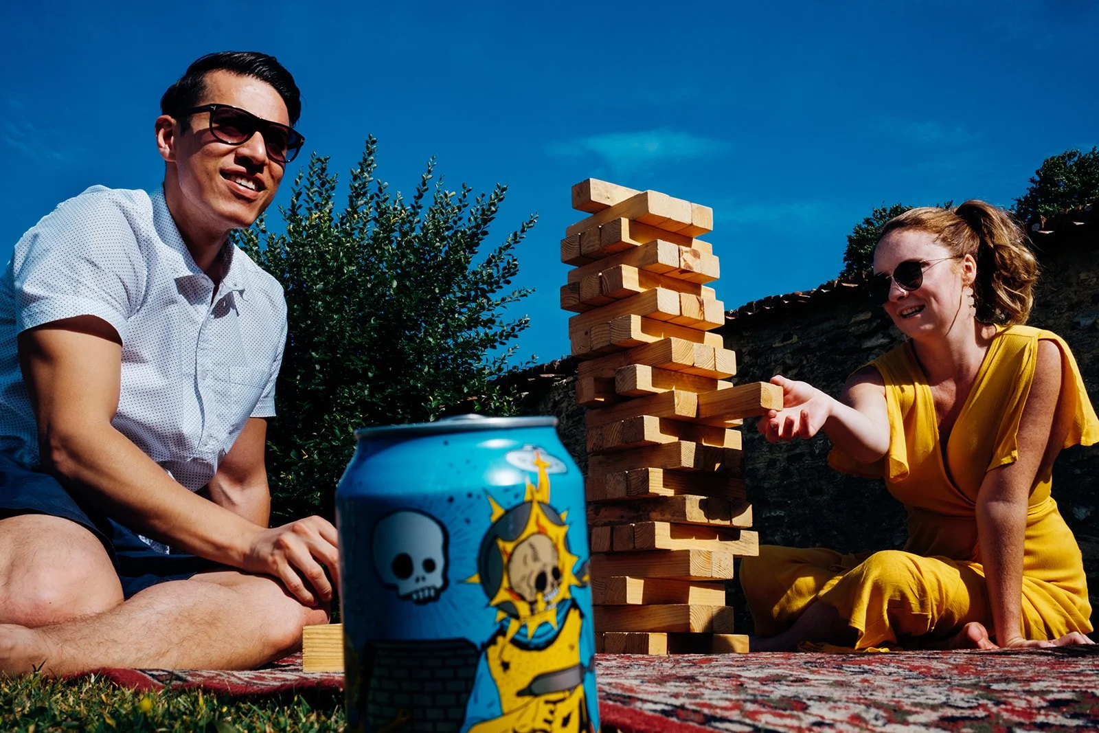 Two people sit on a patterned rug outdoors on a sunny day, playing a large wooden block stacking game, with one person in a yellow outfit carefully pulling out a block while the other watches and smiles. A brightly illustrated blue drinks can sits in