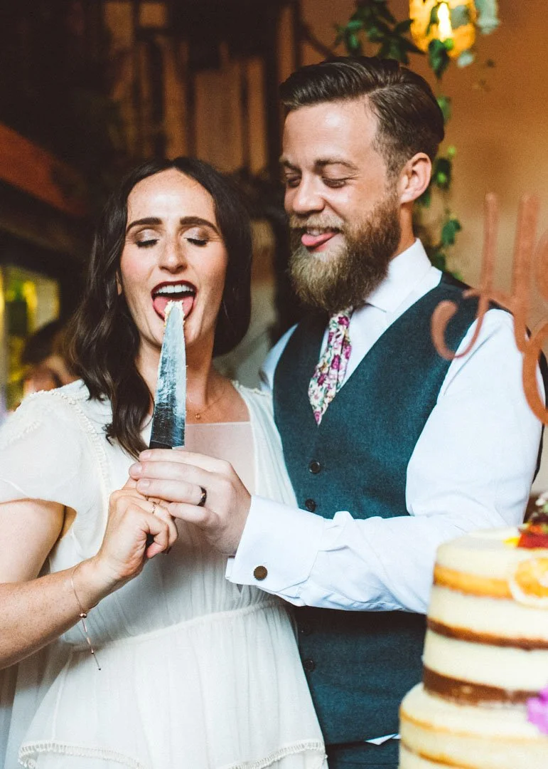 A fun and intimate wedding moment captured by Barbara K Photography, showing a newlywed couple enjoying a playful cake-cutting tradition. The bride, dressed in a soft, vintage-inspired gown, leans in to taste a bit of cake from the knife while her gr