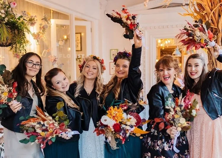 Joyful bridal party celebrating in vibrant autumn colours, featuring glowing, natural makeup by Glamorama Make-up Artist Liverpool. The bridesmaids wear leather jackets and hold rustic floral bouquets, creating a fun, modern twist on traditional wedd