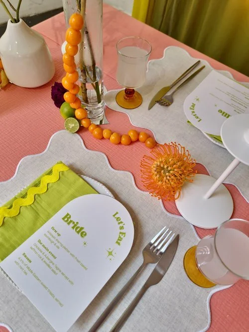A playful and modern wedding tablescape styled by Orange Tree Weddings, featuring bright citrus-inspired details with orange and lime accents, scalloped linens in pink and white, and bold contemporary stationery. The vibrant colour palette adds a fre