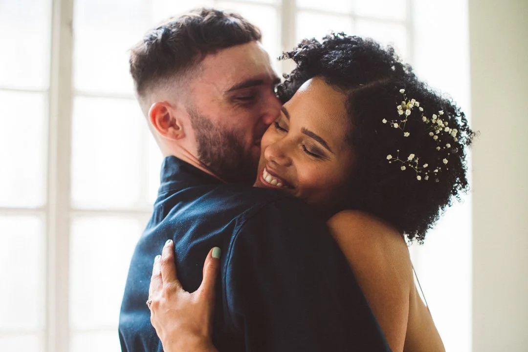 A soft and intimate wedding moment captured by Barbara K Photography, showing a couple wrapped in a warm embrace near a bright window. The bride smiles with her eyes closed, her hair adorned with delicate baby's breath, while the groom holds her clos