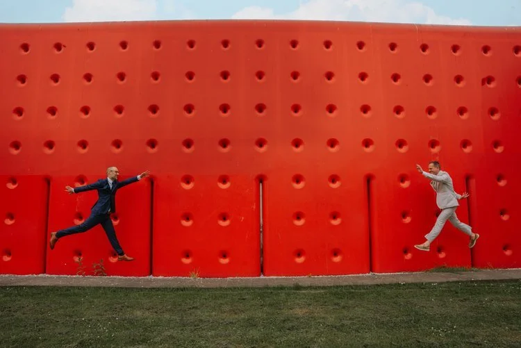A couple leap playfully toward each other in front of a bright red textured wall, captured by Joshua Humphrey Photography. The bold modern backdrop and energetic movement create a fun creative wedding portrait perfect for couples who love vibrant con