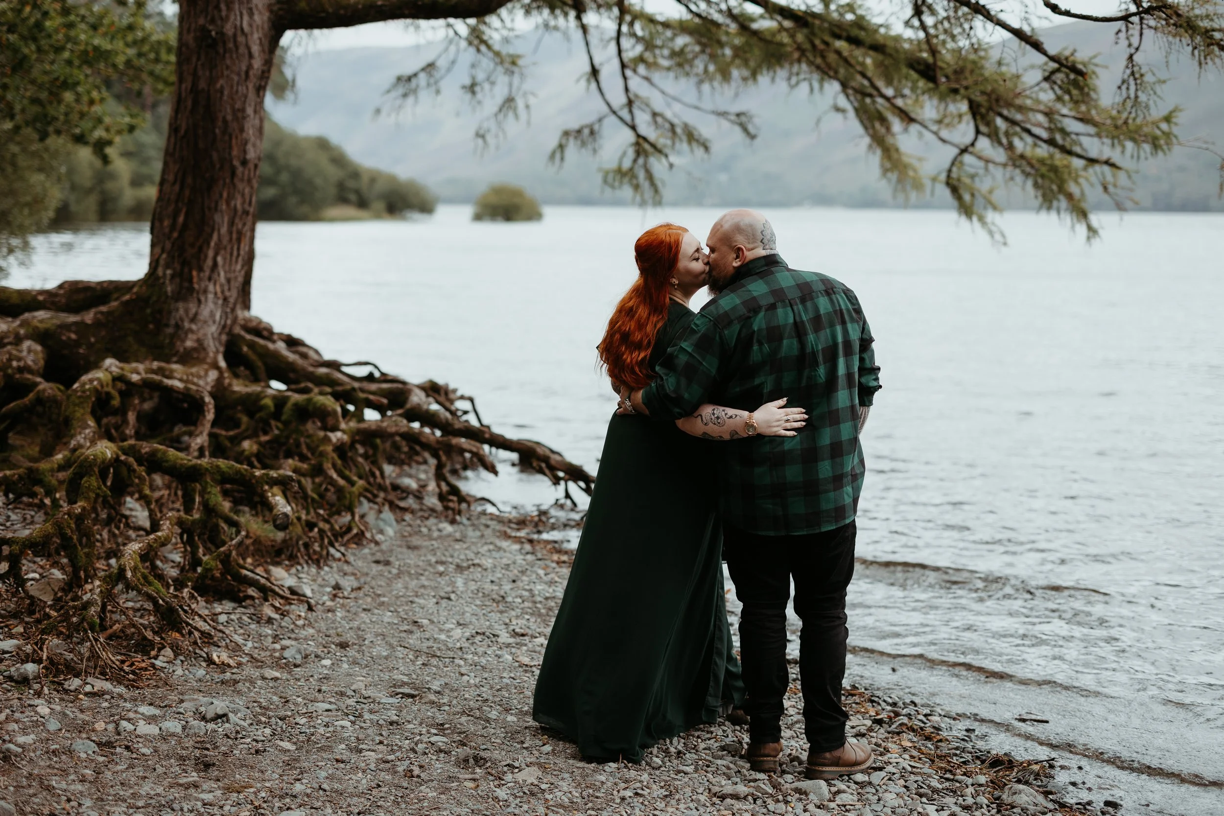 A quiet and romantic lakeside moment captured by Joy Louise Photography, showing the couple sharing a gentle kiss as they stand together on a pebbled shoreline. With towering tree roots twisting along the ground beside them and soft mountains fading 