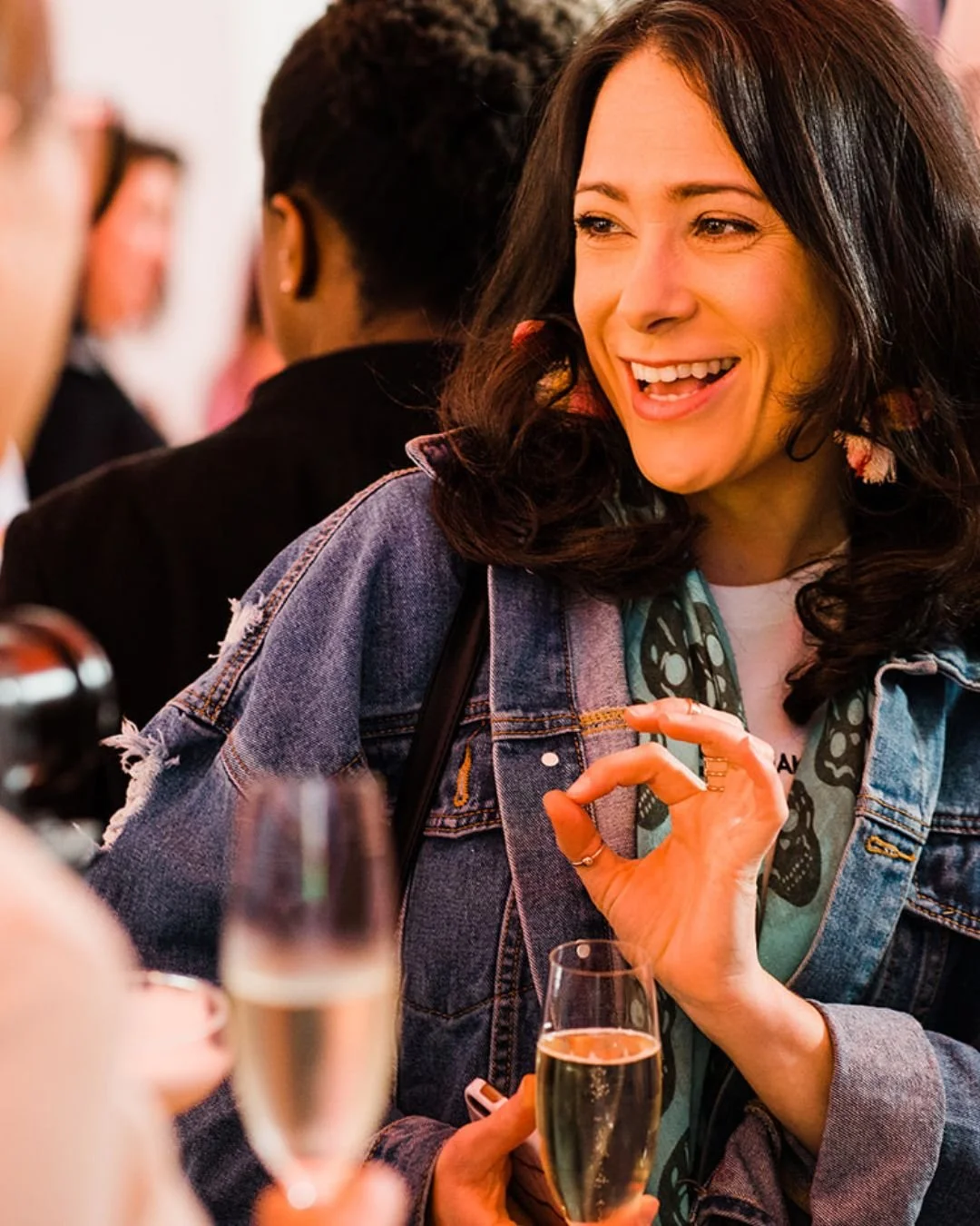 A modern bride-to-be in a denim jacket is drinking champagne at a wedding event in London. She looks happy.