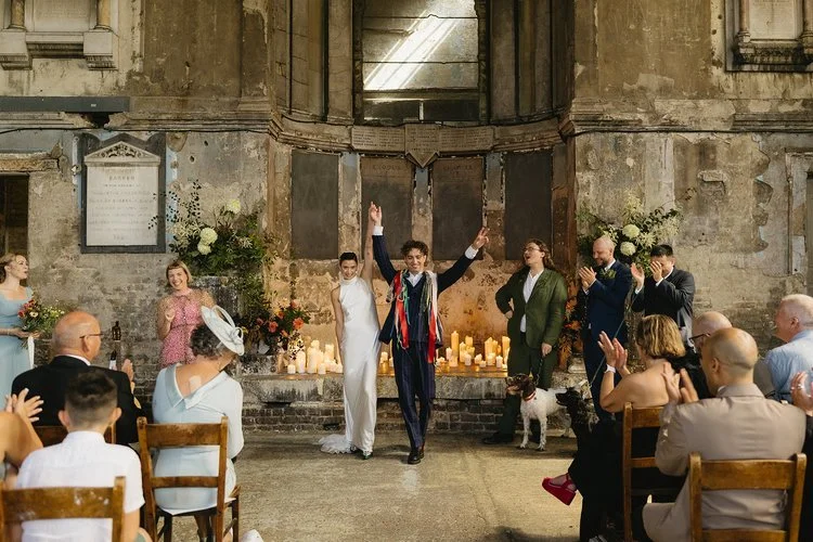 A joyful couple celebrates their marriage in a stunning candlelit ceremony set in an atmospheric, historic venue. Tab Taylor wedding celebrant stands beside them, smiling as guests cheer and applaud. The rustic stone backdrop, glowing candles, and el