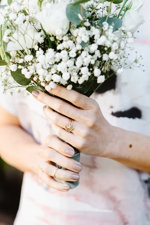 Bride holding a bouquet of white flowers wearing elegant gold stacking rings by Nikki Stark Jewellery. Handcrafted in the UK, these delicate designs add a timeless, minimalist touch to modern bridal style.
