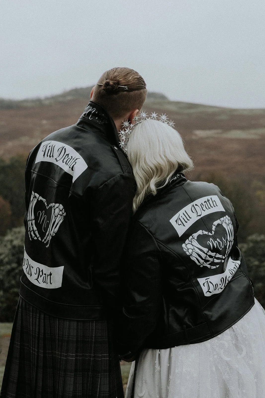 A striking, alternative wedding moment captured outdoors, featuring a couple in custom hand-lettered leather jackets with bold, gothic-style typography and illustrated details. Designed by Toasty Type, the personalised jackets showcase bespoke letter