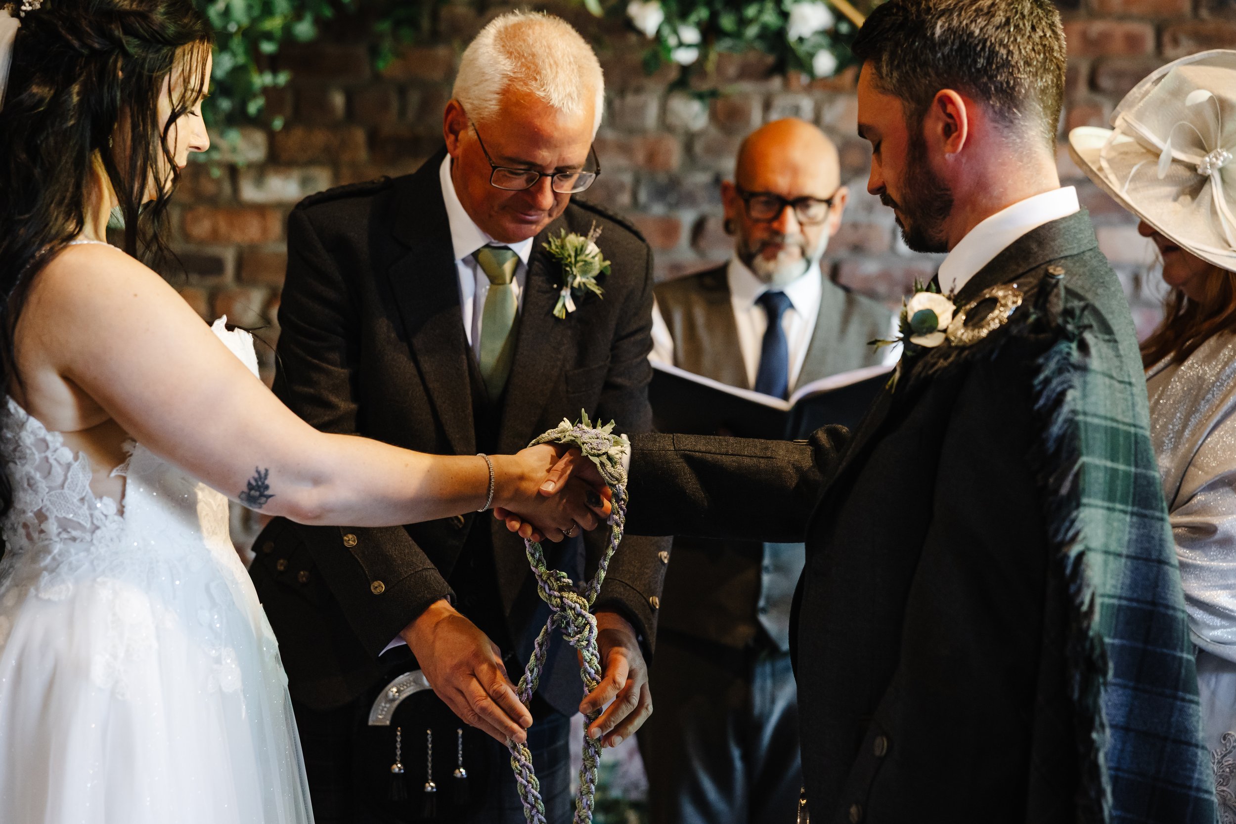 Marie & Oliver's hands are bound with a braided cord during a handfasting ceremony, led by their celebrant. The intimate ritual unfolds against a rustic brick backdrop.