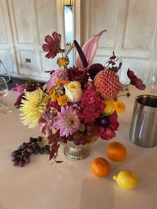 Colourful floral arrangement by Studio Clarisa Floristry featuring dahlias, anthuriums, roses, and cosmos in rich pink, peach, and plum tones. Styled in a vintage vase with fresh citrus fruits and grapes for a luxurious, Mediterranean-inspired weddin