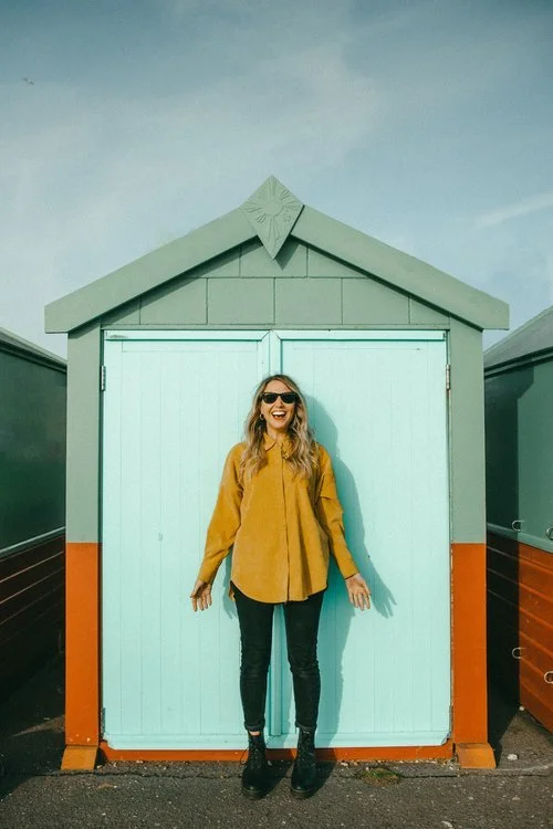 Roxy Celebrates Love standing in front of a brightly coloured beach hut. The hut is painted in a cheerful combination of mint green doors, grey trim, and a rust-orange lower section. Roxy is smiling widely, wearing sunglasses, a mustard-yellow shirt,