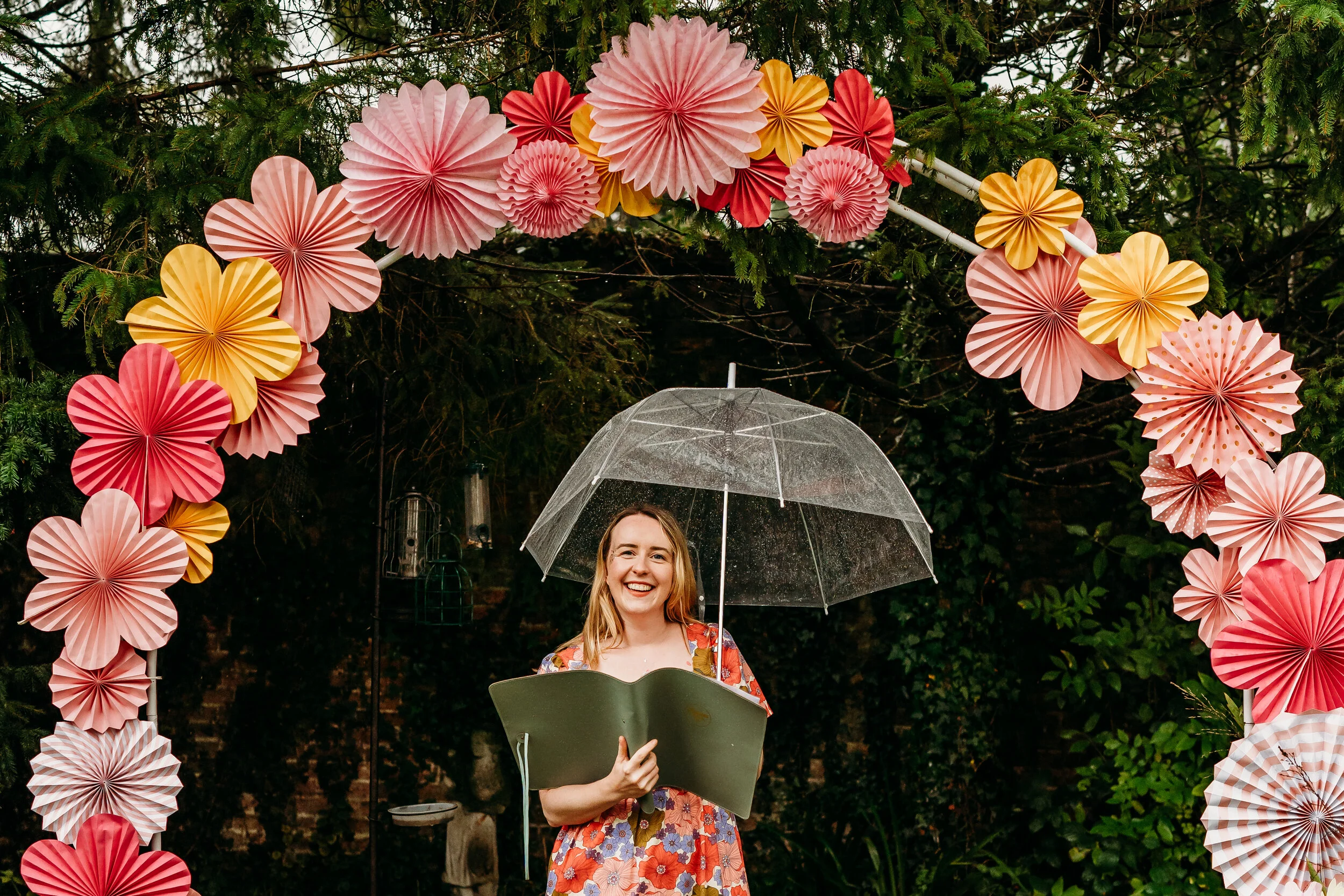 A joyful wedding celebrant, Frances from Frances Celebrates You, stands smiling beneath a colourful paper-flower arch while holding an umbrella during an outdoor ceremony. Surrounded by vibrant pink, red, and yellow decorations, she brings warmth and