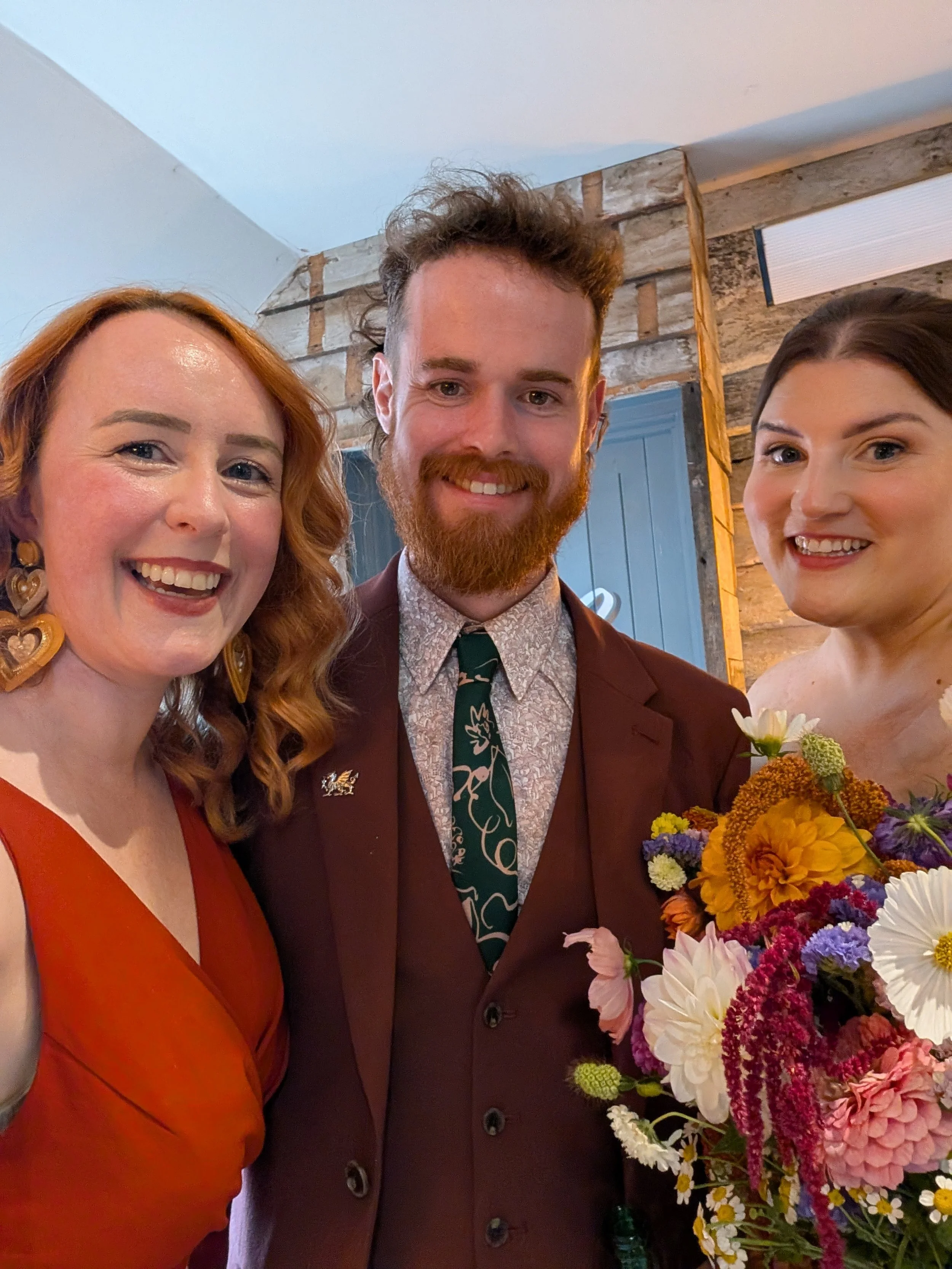 Frances from Frances Celebrates You, a fun and heartfelt UK wedding celebrant, smiling with a newly married couple after an intimate ceremony. The trio stand close together indoors, with the bride holding a bright, seasonal bouquet of colourful flowe