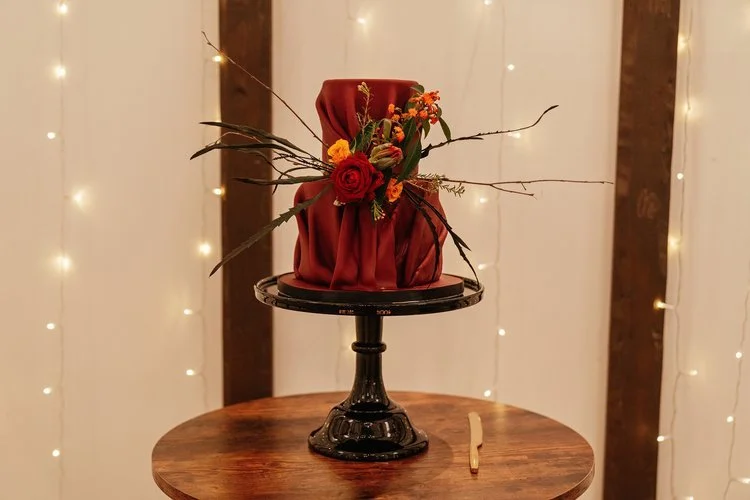 A strikingly elegant creation by Amber’s Cakes and Treats, this unique single-tier wedding cake exudes modern sophistication and autumnal warmth. Draped in deep burgundy fondant that mimics luxurious fabric folds, it’s beautifully accented with a vib