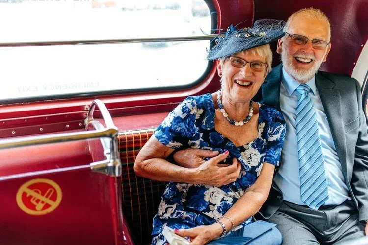 Two wedding guests share a joyful moment while riding on a classic red bus, captured with warmth and humour by Jordanna Marston Photography. Their bright smiles, colourful outfits and the nostalgic bus interior create a fun, heartfelt scene that refl