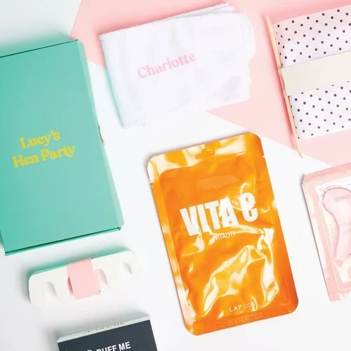 Flat lay of a personalised hen party gift set including a turquoise box with 'Lucy's Hen Party', skincare sachet, towel with name embroidery, and polka dot packaging, created by Melon Teaspoon.