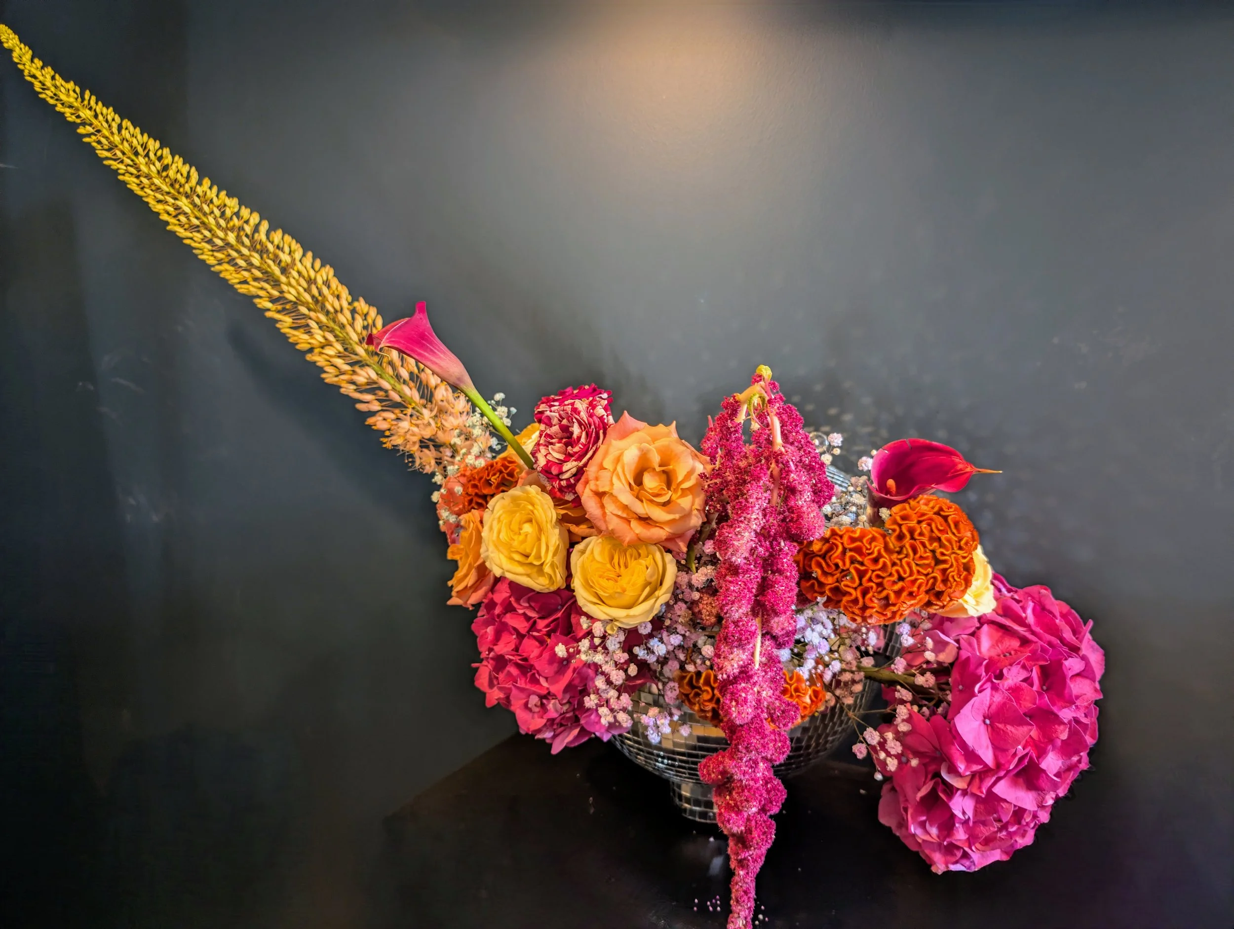 Contemporary floral arrangement by Harlequin Florals featuring a striking design with orange roses, magenta hydrangeas, pink calla lilies, and hanging amaranthus. Set against a dark background, this bold composition highlights modern texture and vibr