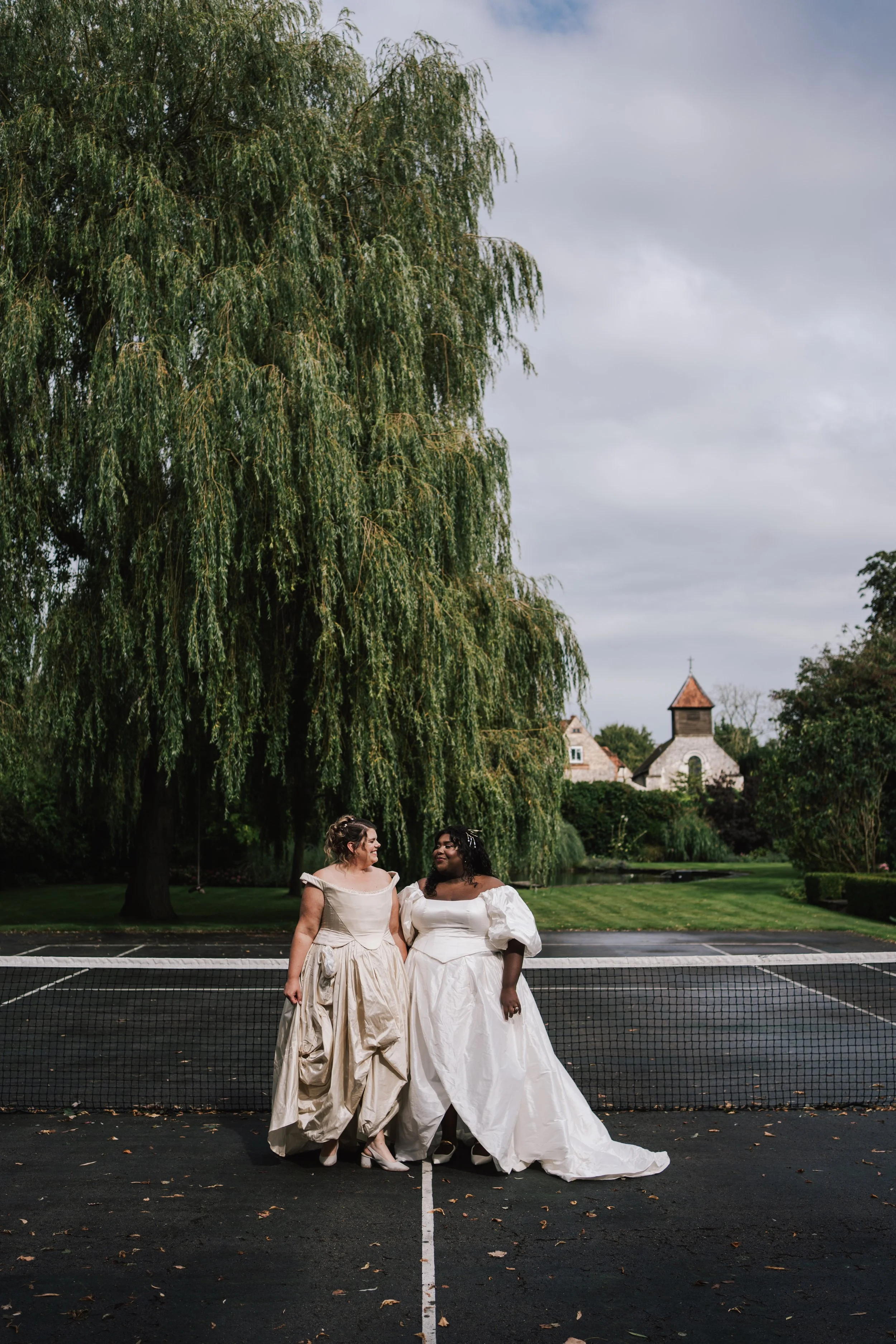 Two brides stand together on an outdoor tennis court, sharing a warm, joyful moment beneath the sweeping branches of a grand willow tree. Their elegant gowns—one with sculptural draping, the other with romantic puffed sleeves—contrast beautifully aga