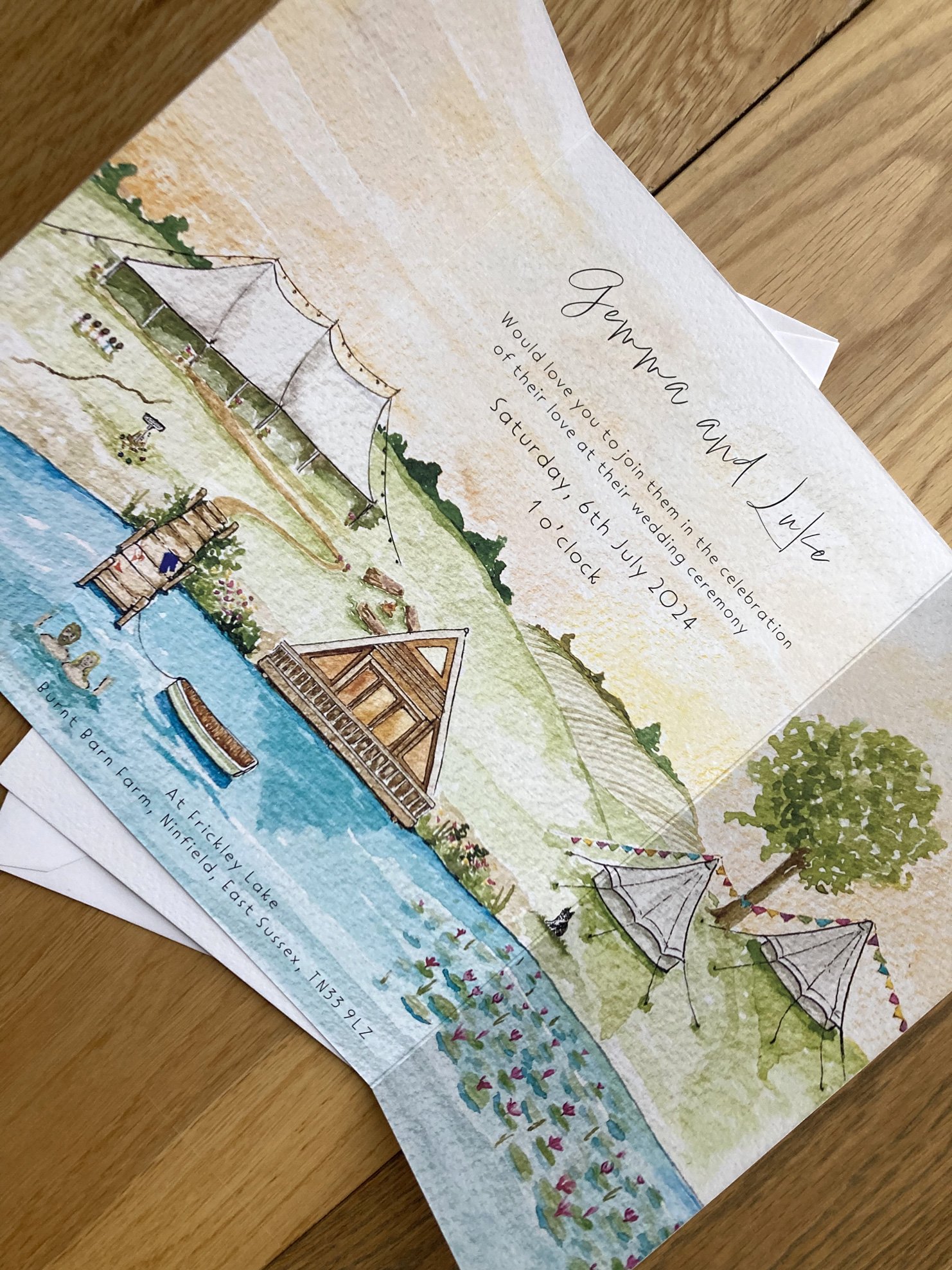 A joyful lakeside wedding invitation by Feather & Ink Stationery featuring a hand painted watercolour scene of Frickley Lake with a wooden boathouse, sailcloth tents, bunting and lily pads printed on textured folded card and styled on warm wooden boa