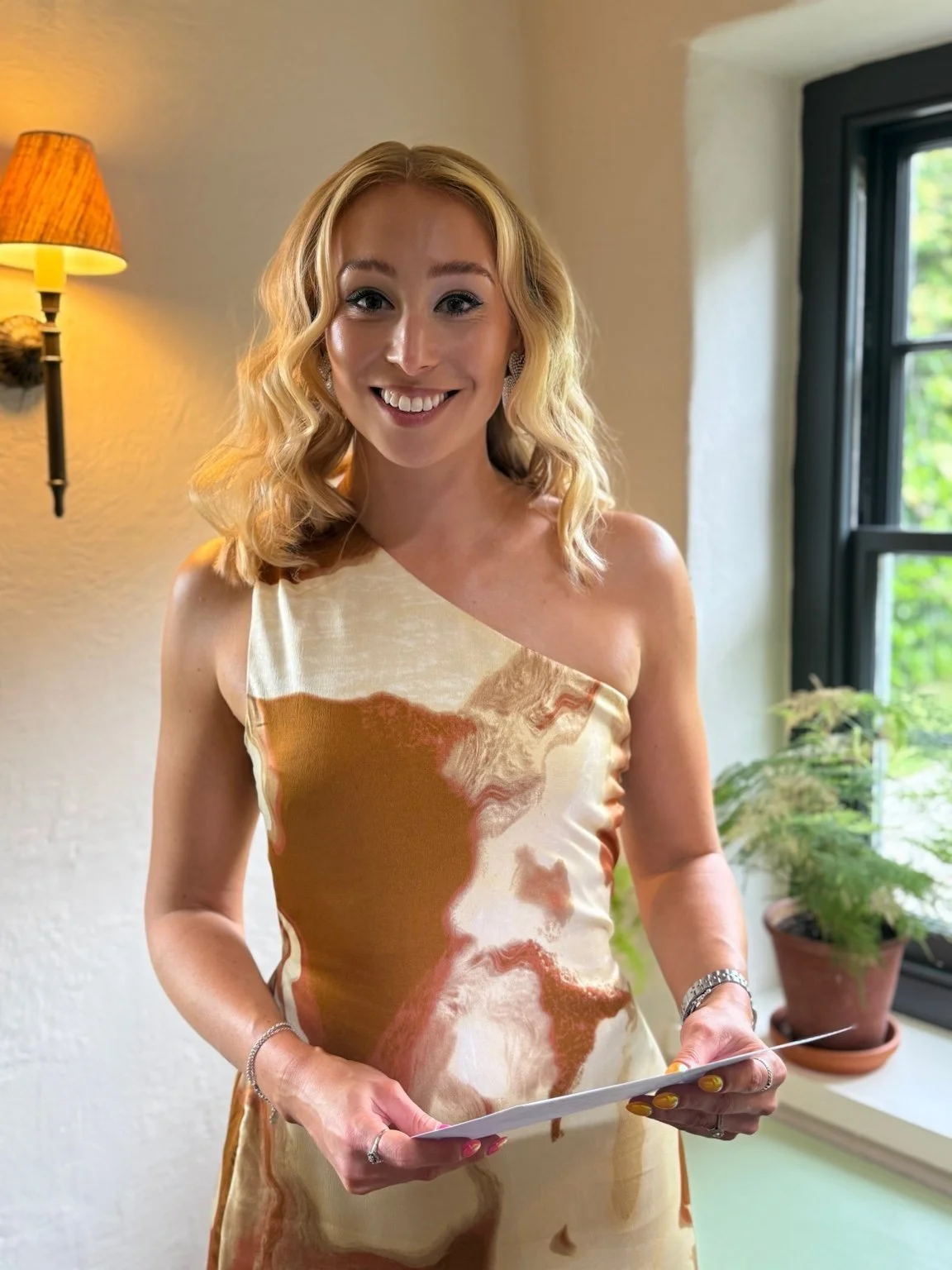 Celebrate with Hayley is smiling at the camera and wearing a beautiful, one shoulder dress with brown and cream patterns.