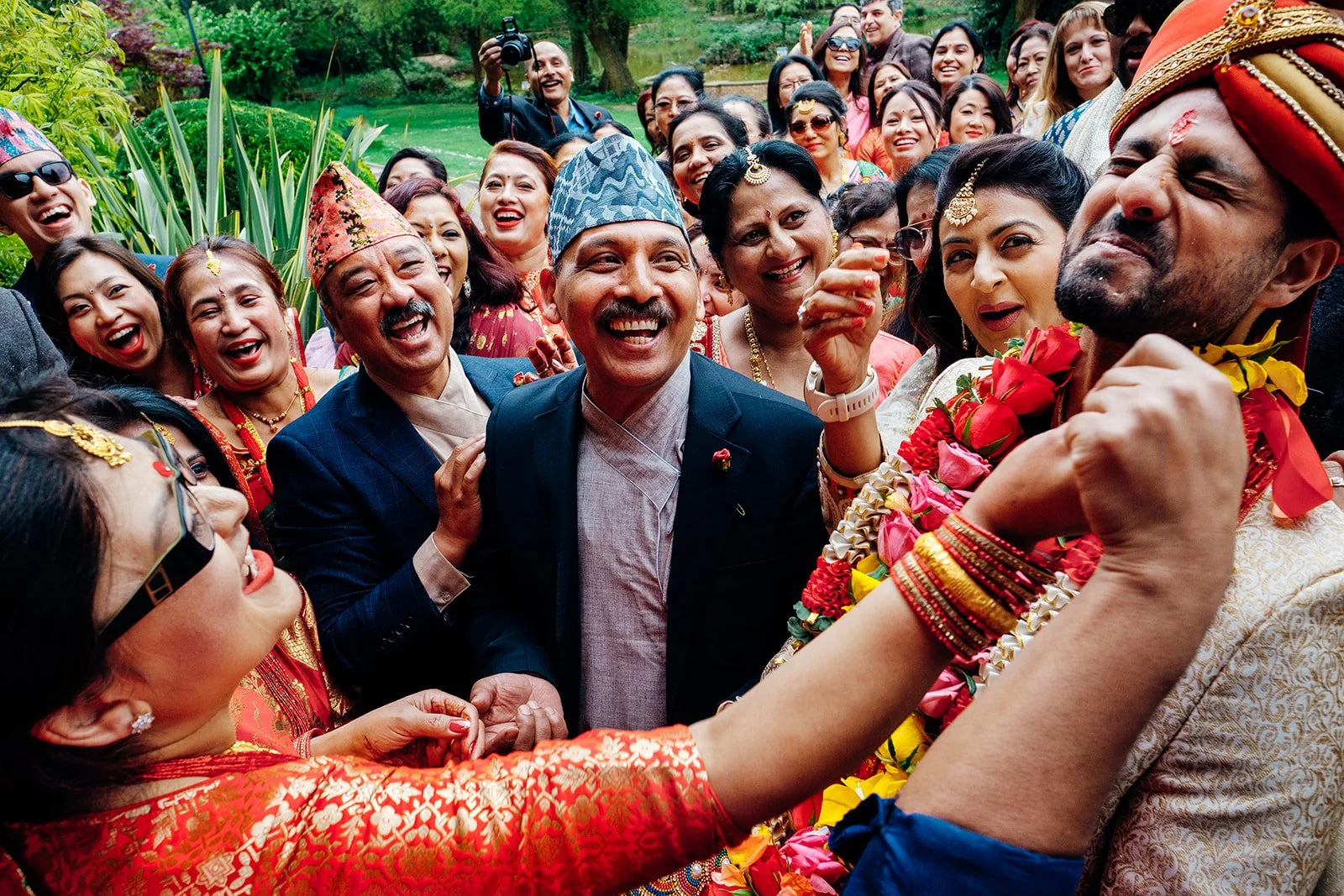 A large group of people dressed in colourful traditional attire are gathered closely together outdoors, smiling and laughing during a lively celebration. Several individuals wear patterned topis and ornate jewellery, while one person on the right, ad