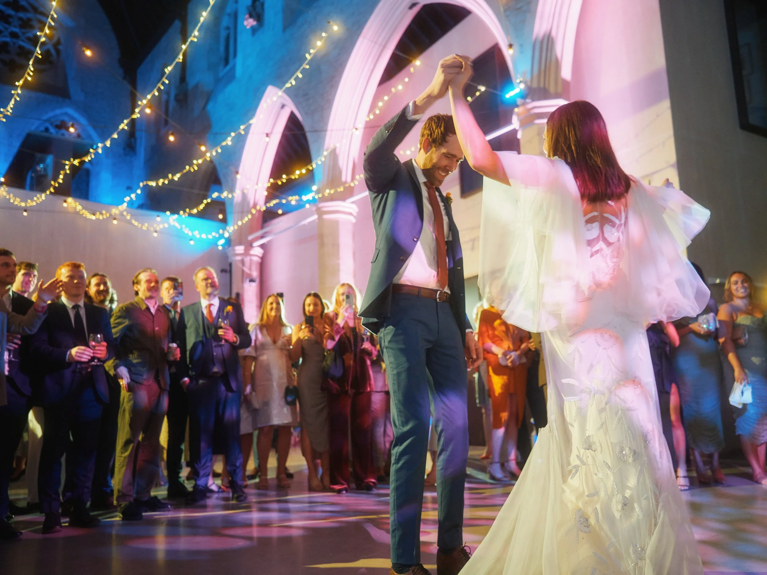 A vibrant and dynamic wedding moment captured by Wayne La Photography, showing the couple sharing their first dance surrounded by friends and family. The groom lifts the bride’s hand confidently as they turn together, illuminated by colourful lightin
