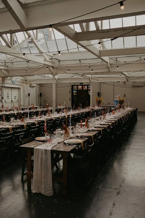 Industrial-style wedding reception by AMW Events, featuring long wooden banquet tables with black chairs, minimalist floral centerpieces, and string lights hanging from exposed white beams.