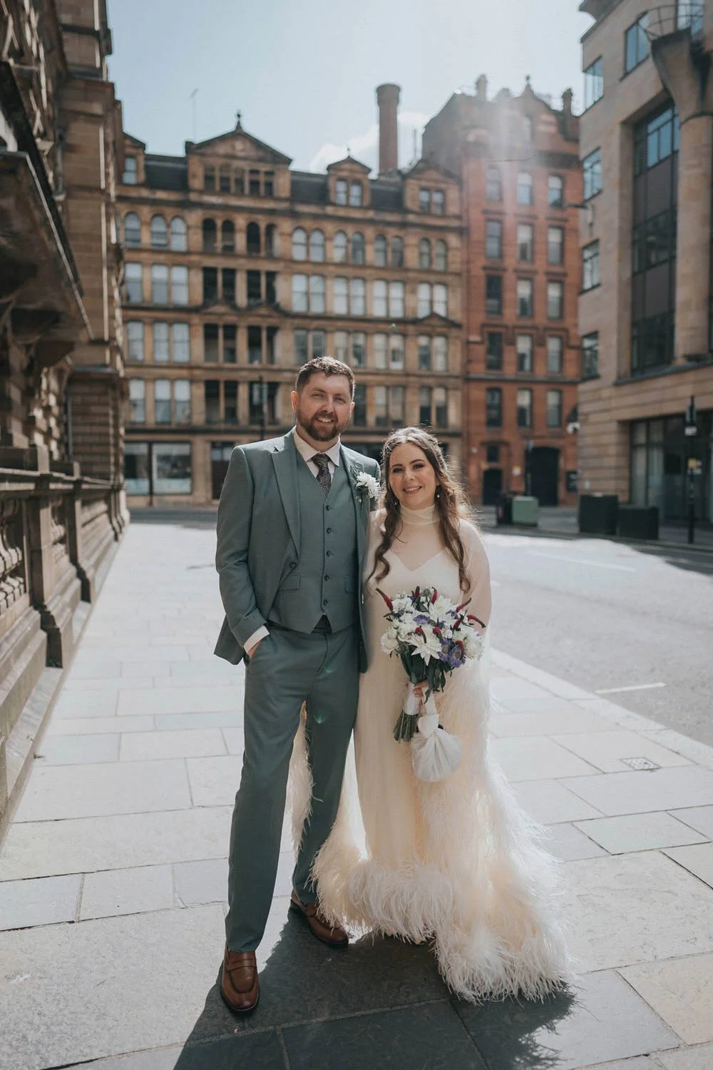 A relaxed city wedding portrait by Good Luck Wolf Photography showing a newlywed couple standing hand in hand on a Glasgow street, with the bride in a long-sleeved feather-trim wedding dress holding a bouquet and the groom in a green three-piece suit
