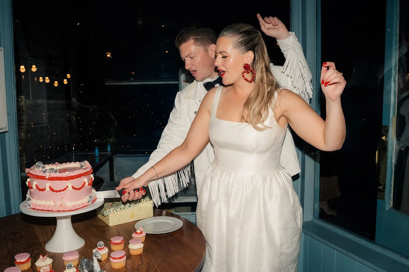 Bride and groom cutting a retro-inspired wedding cake by The Cake Hun, featuring vintage piping, soft pink buttercream and bold red detailing, styled with matching cupcakes. A modern, design-led wedding cake perfect for creative, contemporary wedding