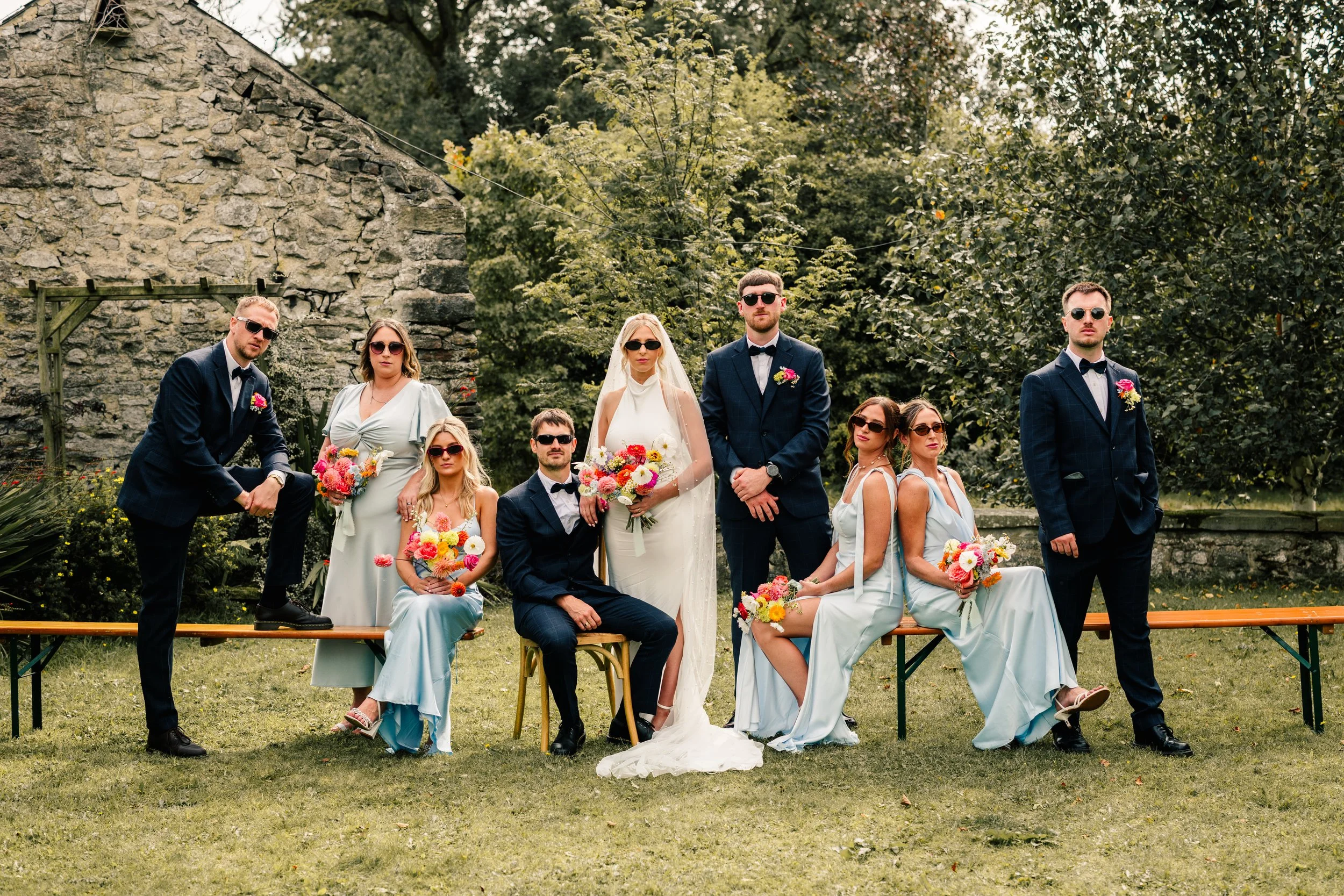 A stylish wedding party poses outdoors in the Peak District, creating a bold editorial group portrait. The bride stands centre-stage in a sleek halter wedding dress and veil, surrounded by bridesmaids in soft blue satin dresses and groomsmen in navy 