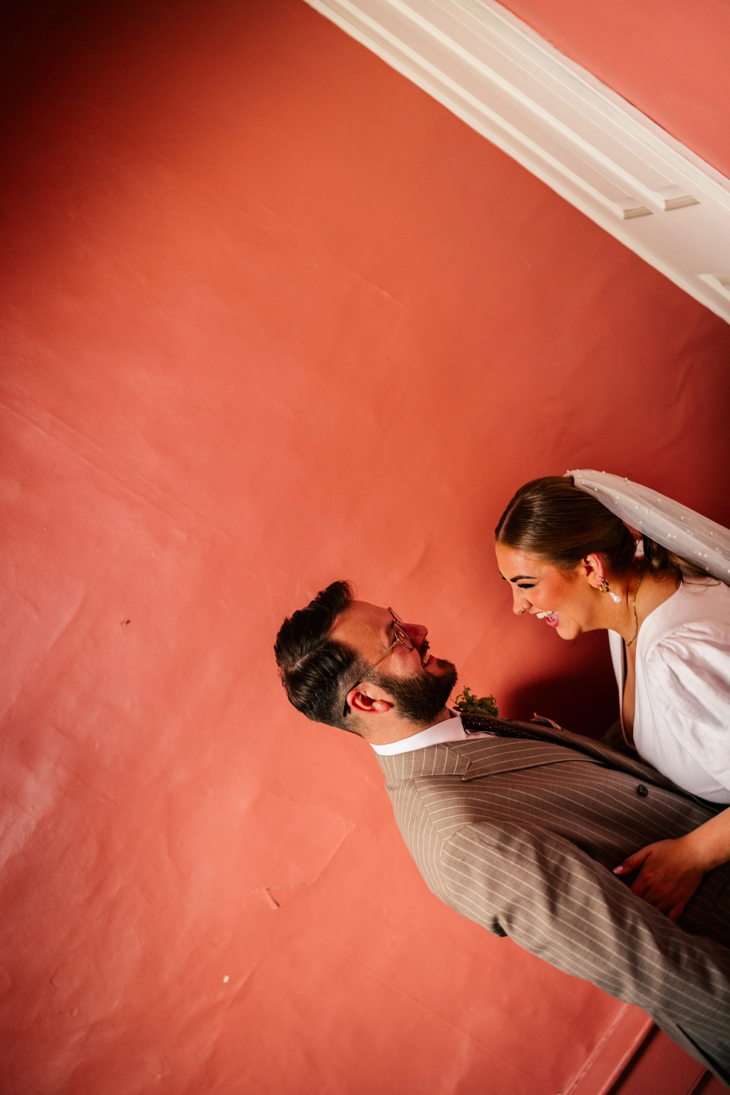A joyful newly married couple share a candid moment inside a warm, terracotta-painted room during their Winchester townhouse wedding. The groom in a tailored pinstripe suit leans in laughing with the bride, who wears a modern white dress with puff sl