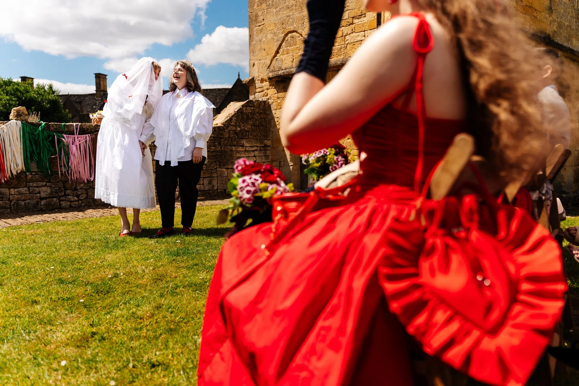 Two people stand together on a grassy outdoor area beside an old stone building, with one wearing a long white outfit and veil decorated with small red bows and the other wearing a white shirt and black trousers, both laughing as they hold hands. In 