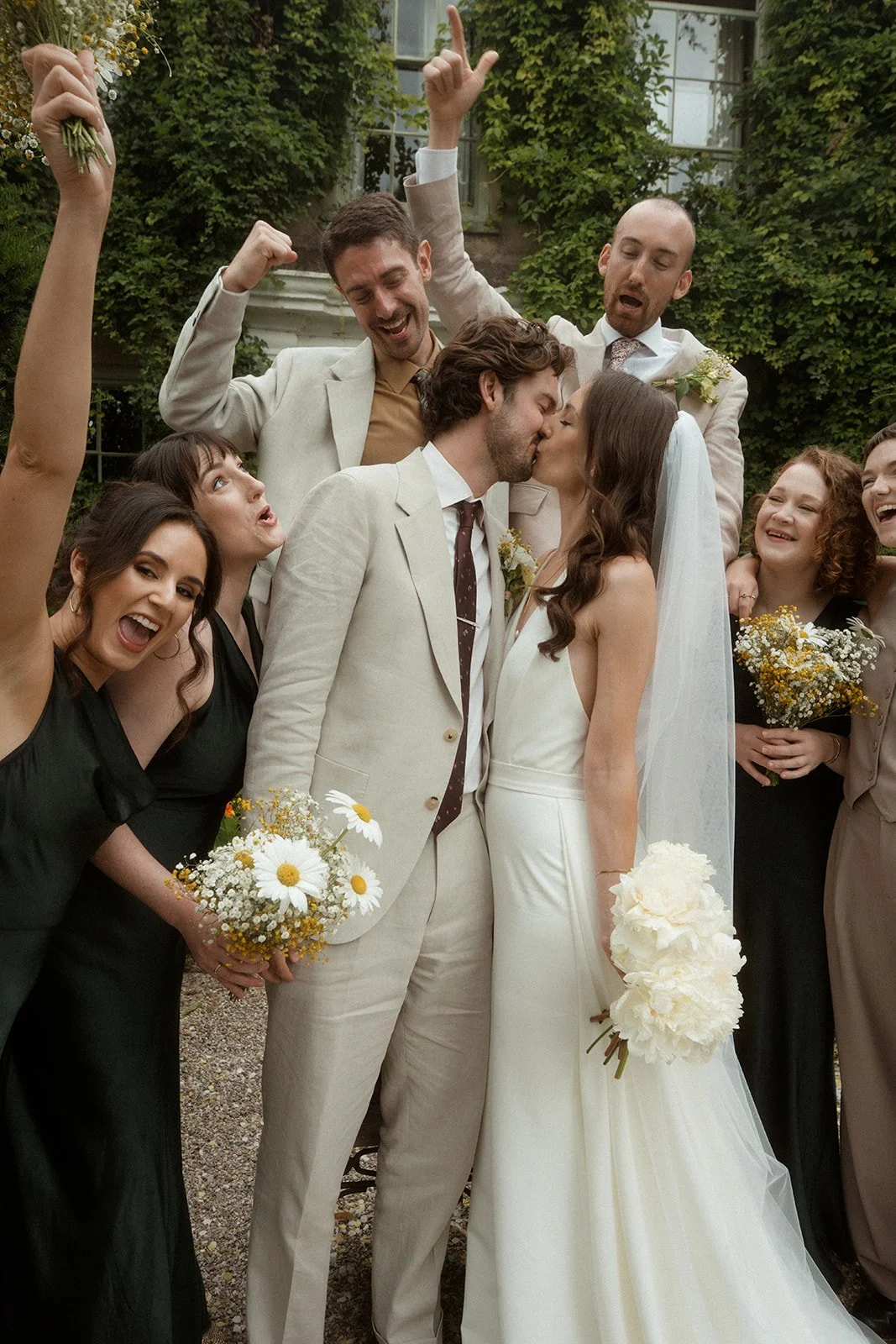 This wedding group photo features a bride and groom kissing in front of a historic ivy covered building while their bridal party surrounds them cheering, with the bride holding white peonies and the groom wearing a light beige suit as bridesmaids and