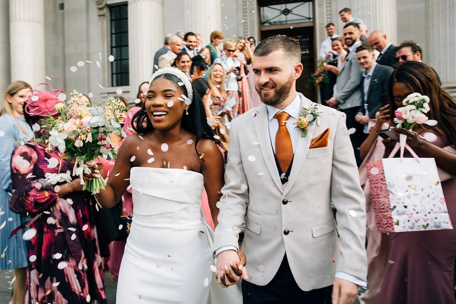 A joyful rush of movement as a newly married couple walks through a cascade of soft white confetti, surrounded by cheering guests. They’re grinning, hand in hand, totally swept up in the celebration. The bride wears a sleek strapless white gown with 