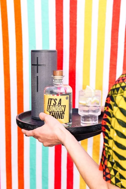 A person holds a black serving tray with a bottle of Rythmik Cocktails’ It’s A London Thing gin cocktail, a glass with lemon garnish, and a portable speaker in front of a bright, striped backdrop. The colourful setting captures a fun, music-filled vi