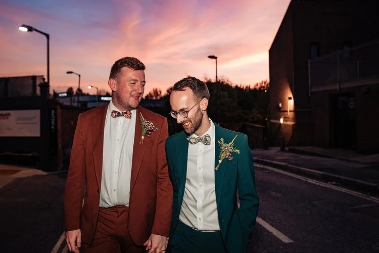A vibrant and contemporary wedding portrait captured by Natalia Case Photography, showing the couple walking hand in hand beneath a stunning sunset sky. One groom wears a rich rust suit and the other a deep teal, their complementary colours creating 