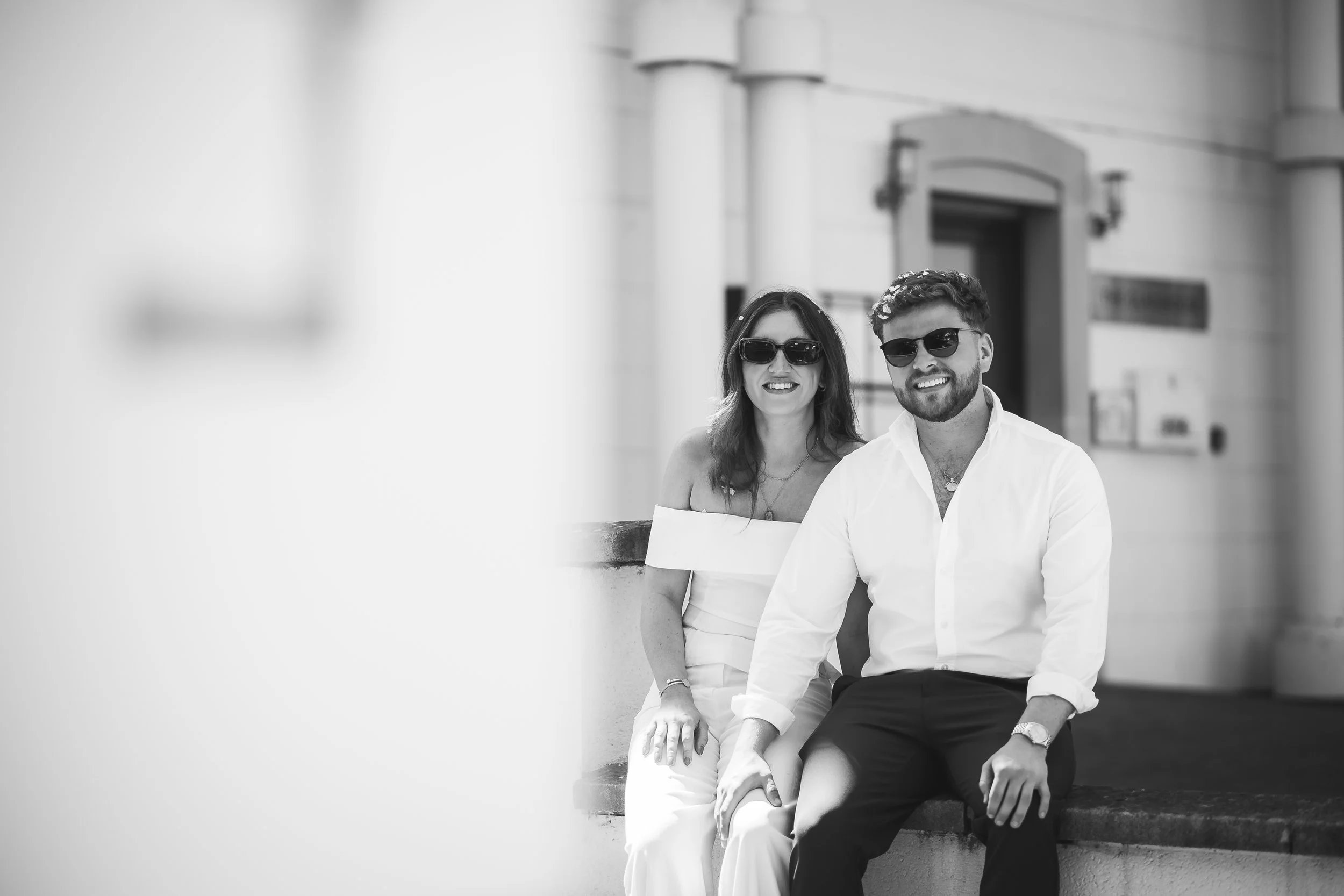 A stylish black-and-white engagement photo captured by Gemma Gaskins Photography, featuring a couple sitting together outdoors in natural light. Their relaxed smiles, matching sunglasses, and modern outfits create a cool, contemporary feel—perfect fo