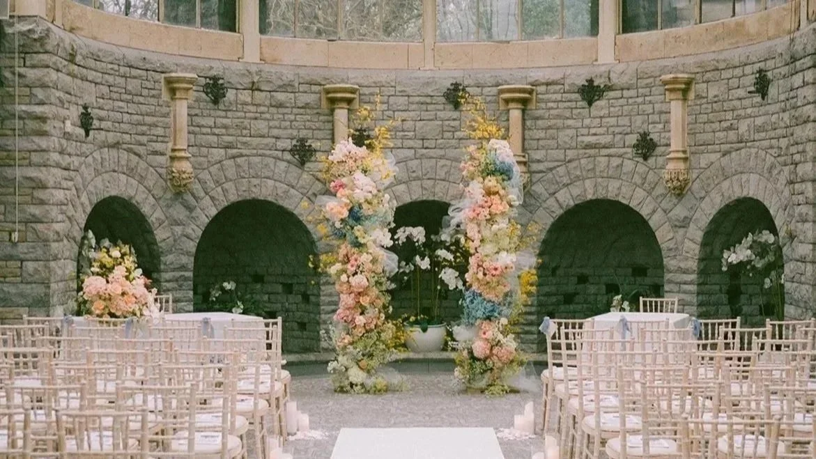 Elegant wedding ceremony setup by AMW Events in a grand glass-domed venue with pastel floral arrangements, white aisle runner, and candlelit decor.
