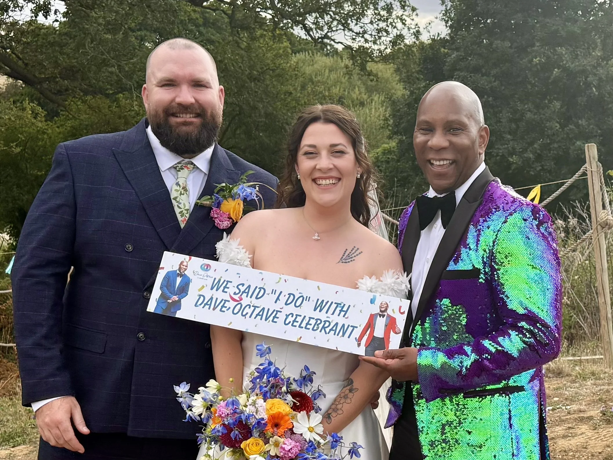 Dave Octave Celebrant in a colourful sequinned blazer, with a bride and groom, holding a sign that reads 'We said "I do" with Dave Octave Celebrant". 