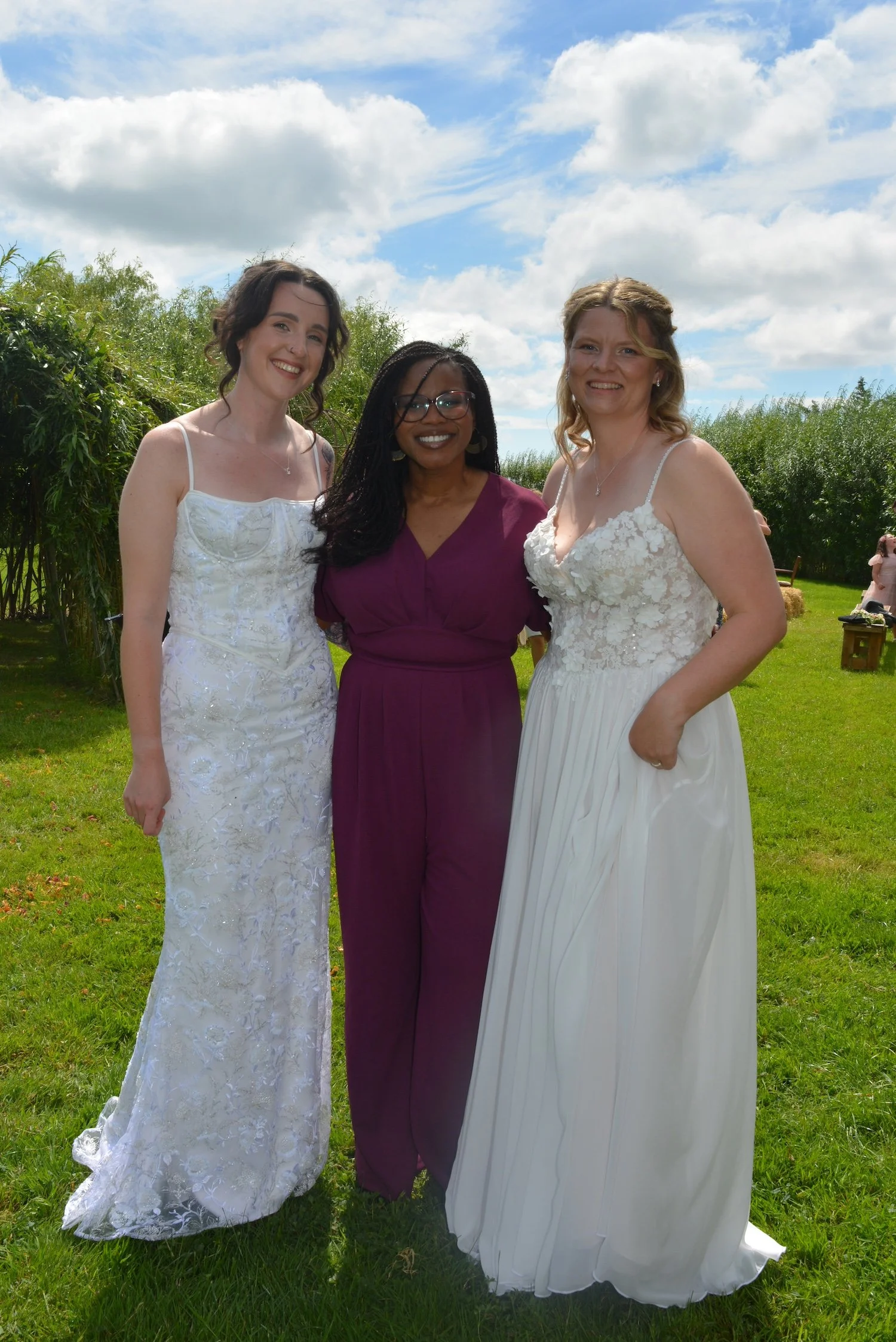Deyi from Gather & Tell Ce;ebrations is standing in the middle of 2 brides at an outdoor wedding ceremony.